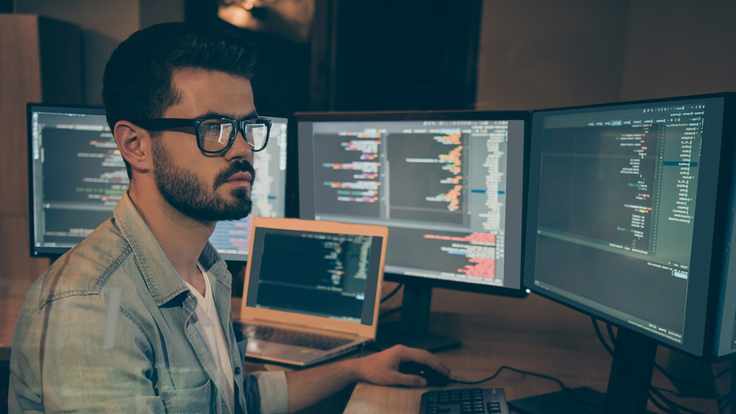 Un joven frente a un grupo de pantallas ejerciendo sus conocimientos en sistemas computacionales después de cursar una ingeniería en línea.